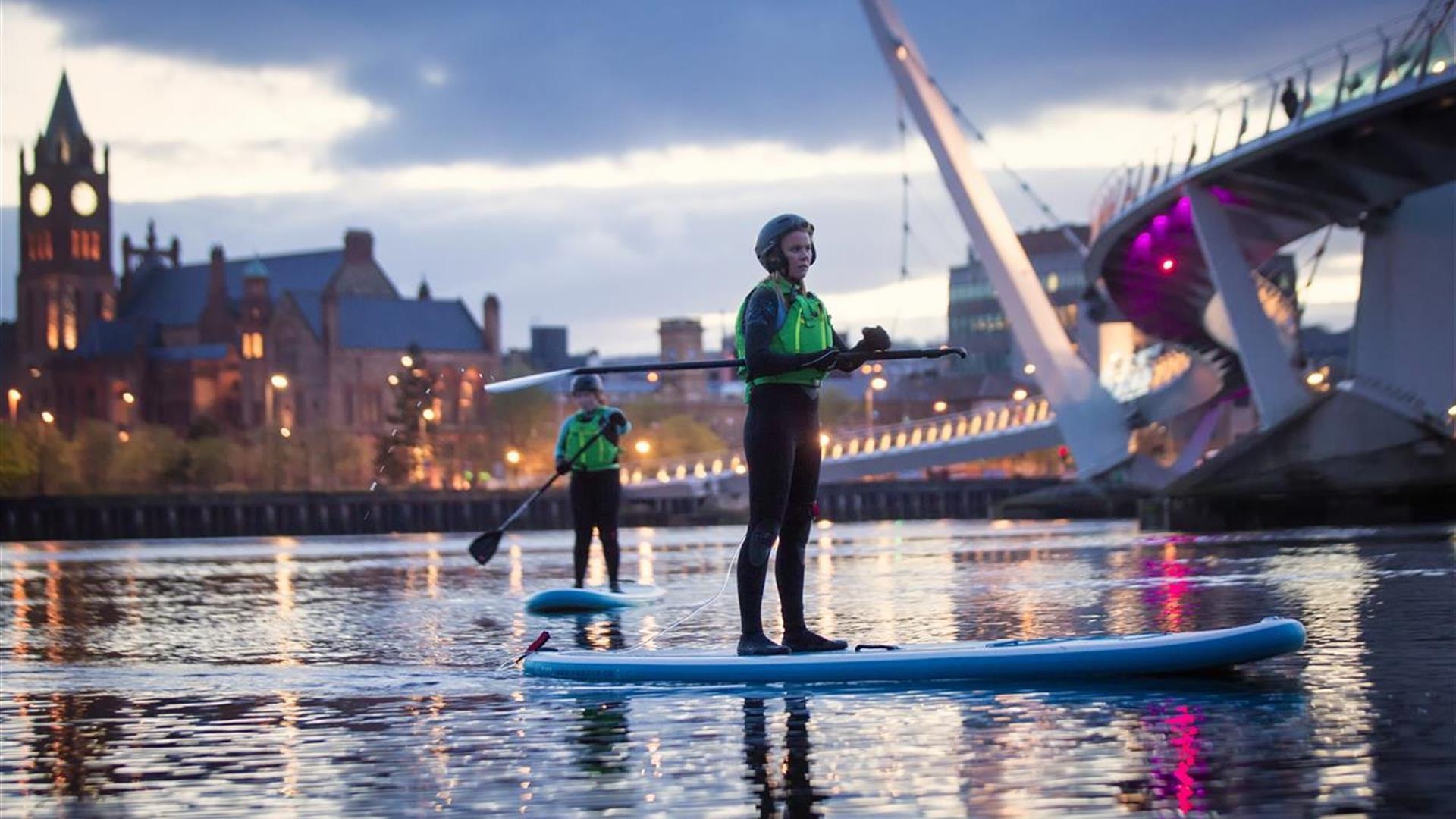 Paddleboarders on the River Foyle at dusk, with the Guildhall and the Peace Bridge behind