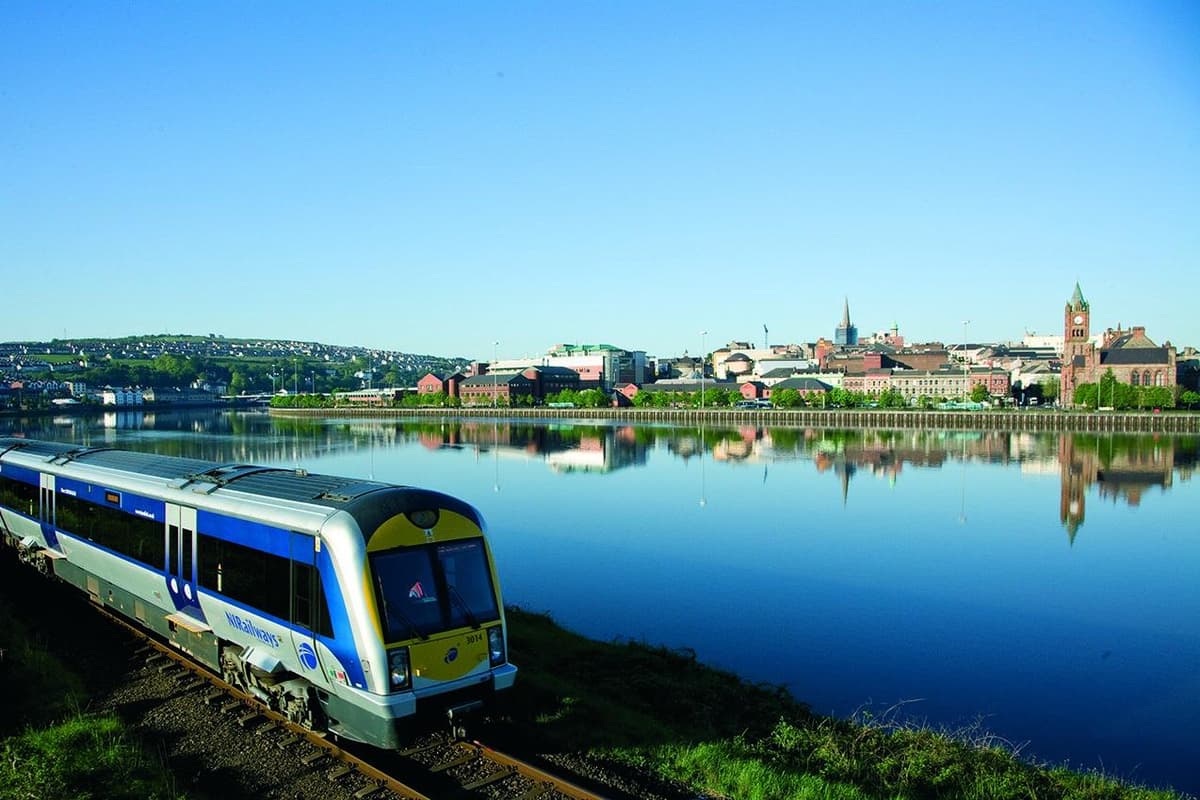 An NI Railways train beside the River Foyle with the Derry skyline behind
